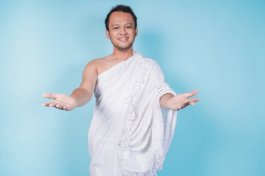 Zarif, Asyalı, ihram giyen genç Müslüman bir adam davet ediyor ya da jest yapmaya gidiyor, mavi arka planla izole edilmiş. Hac kavramı.