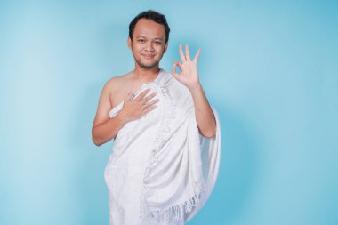 İhram giyen neşeli, Asyalı genç Müslüman adam mavi arka planla izole edilmiş parmaklarını kullanarak tamam işareti gösteriyor. Hac kavramı.