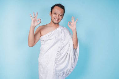 İhram giyen neşeli, Asyalı genç Müslüman adam mavi arka planla izole edilmiş parmaklarını kullanarak tamam işareti gösteriyor. Hac kavramı.