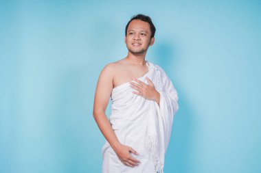 İhram takan mutlu Asyalı genç Müslüman adam izole edilmiş mavi arka planda el ele poz veriyor. Hac kavramı.