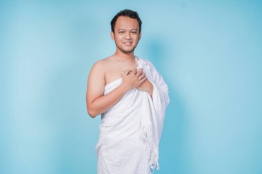 Mutlu, düşünceli, genç, Asyalı Müslüman adam el ele tutuşup İhram giyip izole edilmiş mavi arka plan takıyor. Hac kavramı.