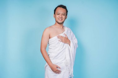 İhram takan mutlu Asyalı genç Müslüman adam izole edilmiş mavi arka planda el ele poz veriyor. Hac kavramı.