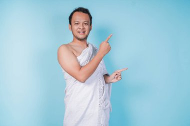 İhram giyen çekici genç Asyalı Müslüman adam yanındaki fotokopi alanını işaret ediyor, mavi arka planla izole edilmiş. Hac kavramı.