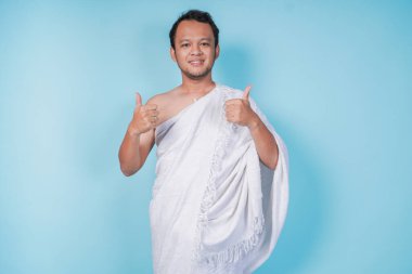 Heyecanlı, Asyalı, ihram giyen genç Müslüman adam mavi arka planla izole edilmiş bir şekilde el hareketi yapıyor. Hac kavramı.