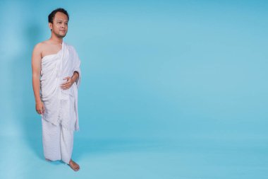 İhram giyen yakışıklı Asyalı genç Müslüman adam mavi arka planla izole edilmiş bir şekilde yürüyor. Hac kavramı.