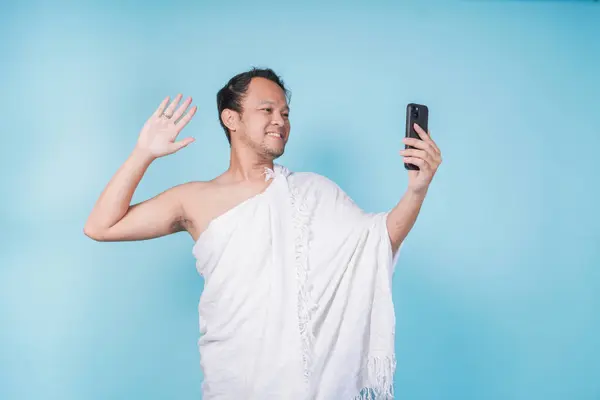 stock image Handsome Asian Muslim man wearing ihram doing video call and waving hello gesture, isolated blue background. Hajj concept.