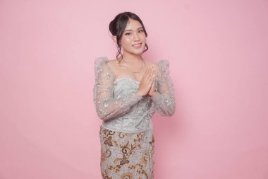 Modern kebaya elbisesi giyen zarif genç Endonezyalı kadın selamlama ya da namaste pembe arka planda izole edilmiş..