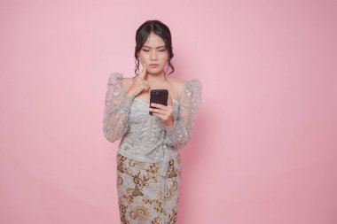 Modern kebaya takan düşünceli genç Endonezyalı kadın, izole edilmiş pembe arka plan üzerinde düşünürken elinde akıllı telefon tutuyor..