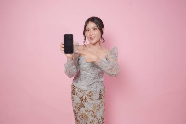 Modern kebaya giysili gülümseyen Endonezyalı kadın akıllı telefonunda yeşil ekran veya fotokopi alanı gösteriyor, izole edilmiş pembe arka plan.