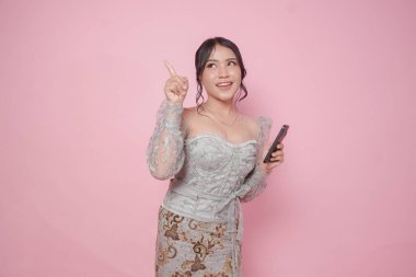 Modern kebaya elbiseler giyen neşeli Asyalı kadın elinde pembe arka planla izole edilmiş akıllı bir telefonla fotokopi odasını işaret ediyor..