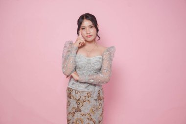 Modern kebaya elbisesi giyen düşünceli Asyalı kadın başını eğmiş, pembe arka planla izole edilmiş..