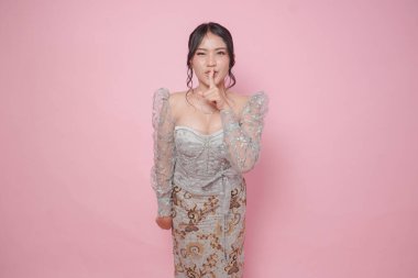 Modern kebaya elbisesi giyen genç Asyalı kadın parmağını dudaklarının önüne koyuyor ve işaret ediyor: 