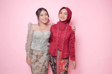 İki neşeli Asyalı genç kadın modern kebaya giyip birbirlerine sarılıyorlar. Pembe arka planda soyutlanmış arkadaşça bir jest yapıyorlar..