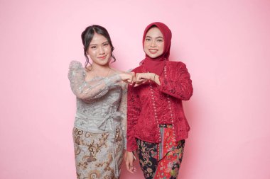Modern kebaya takmış gülümseyen Asyalı genç kadınlar pembe arka planda izole edilmiş yumruk tokuşturma işareti gösteriyorlar..