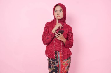 Modern kebaya ve tesettüre bürünmüş düşünceli genç Endonezyalı kadın akıllı telefonu tutarken soyutlanmış pembe arka plan üzerinde çok düşünüyor..