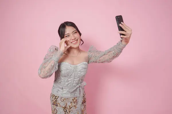 stock image Lovely Indonesian woman in modern kebaya posing for a selfie doing love shape on cheek, isolated by pink background.