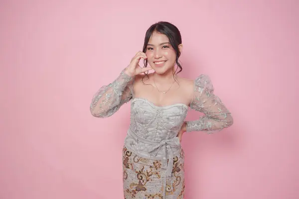 stock image Lovely young Asian woman wearing modern kebaya dress making heart shape gesture on cheek, isolated by pink background.