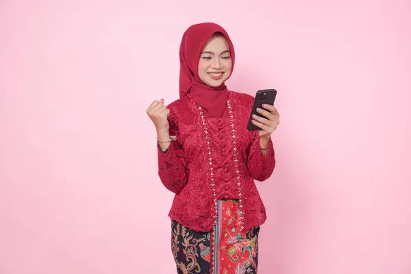 stock image Young Asian woman wearing modern kebaya dress and hijab holding smartphone with a happy successful expression, isolated by pink background.