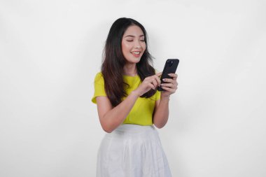 Sarı tişörtlü, beyaz etekli güzel bir Asyalı kadın beyaz arka planla izole edilmiş, geniş bir gülümsemeyle akıllı telefonuna bakıyor..