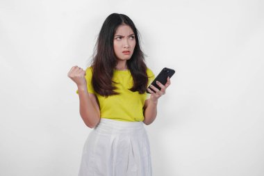 Sarı tişörtlü üzgün bir Asyalı kadın elinde siyah bir cep telefonu tutuyor ve kızgın görünüyor. Suratı asık bir şekilde telefona bakıyor..