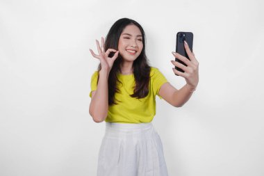 Sarı tişörtlü mutlu Asyalı kadın cep telefonuyla selfie çekiyor. Gülümsüyor ve izole edilmiş beyaz arka planda iyi bir parmak hareketi gösteriyor..