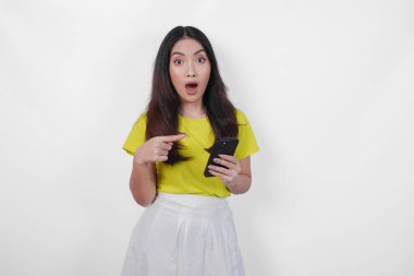 Sarı tişörtlü genç Asyalı kadın, izole edilmiş beyaz arka planda cep telefonunu işaret ediyor..