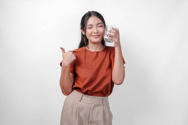 Turuncu gömlekli gülümseyen kadın elinde bir bardak su tutuyor ve başparmağını kaldırıyor. Pozitiflik ve sağlık bilinci kavramı, kadın susuz kalmamanın önemini destekliyor..