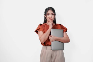 Kahverengi tişört giyen düşünceli genç Asyalı kadın dizüstü bilgisayarını tutuyor ve ciddi bir ifadeyle düşünüyor, beyaz arka planla izole edilmiş..