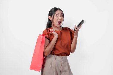 Kahverengi tişört giyen genç Asyalı kadın elinde bir alışveriş çantası ve beyaz arka planla izole edilmiş bir cep telefonu tutuyor..