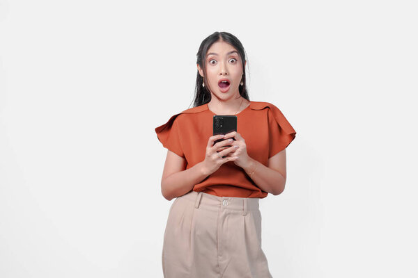 Shocked young Asian woman in brown shirt holding a smartphone with a surprised expression on her face, isolated by white background. — Stock Image Shocked Young Asian Woman Brown Shirt Holding Smartphone Surprised Expression Stock Picture