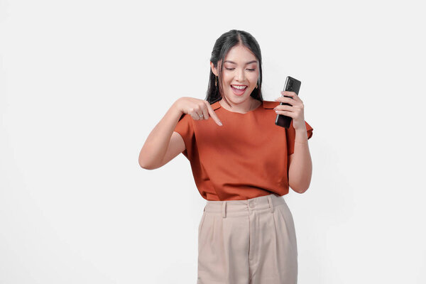 Joyful young Asian woman in brown shirt smiling and holding smartphone while pointing to the copy space downward, isolated white background. — Stock Image Joyful Young Asian Woman Brown Shirt Smiling Holding Smartphone While Royalty Free Stock Photos