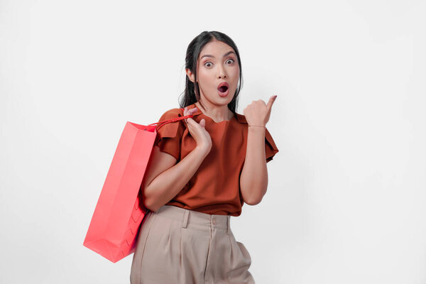 Surprised young Asian woman in brown shirt holding a shopping bag while pointing to the copy space beside her, isolated white background. — Stock Image Surprised Young Asian Woman Brown Shirt Holding Shopping Bag While Stock Image