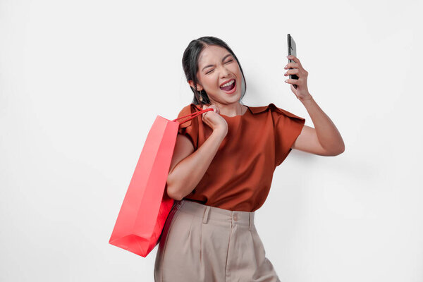 Cheerful young Asian woman in a brown shirt holding a red shopping bag smartphone with happy successful expression, isolated by white background. — Stock Image Cheerful Young Asian Woman Brown Shirt Holding Red Shopping Bag Stock Photo
