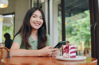 Bir kadın önünde bir pastayla bir masada oturuyor. Bir cep telefonu tutuyor ve kameraya gülümsüyor..