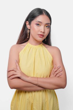 Sarı elbiseli, kendine güvenen genç Asyalı kadın kollarını kavuşturmuş, beyaz arka planda izole bir şekilde poz veriyor..
