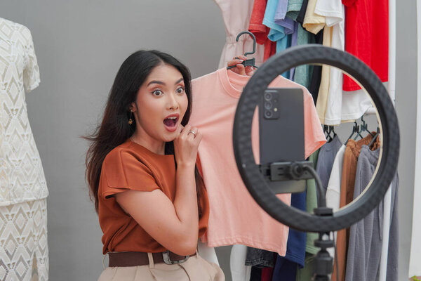Young Asian woman presenting peach colored shirt to the live shopping audience with surprised expression. — Stock Image Young Asian Woman Presenting Peach Colored Shirt Live Shopping Audience Royalty Free Stock Images