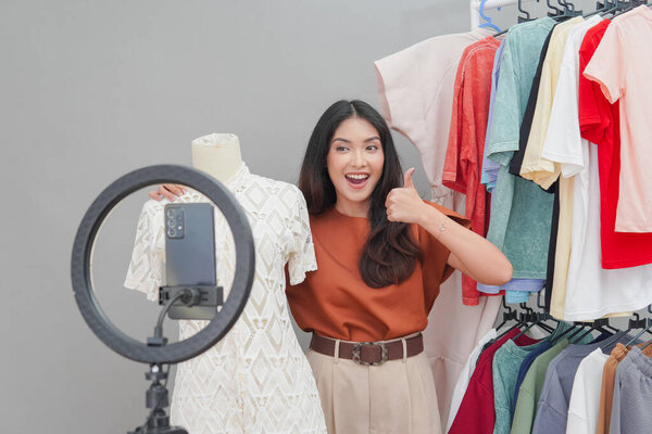 Beautiful Asian woman is holding a mannequin with white dress in front of a rack of clothes while showing thumb up gesture. Live shopping concept. — Stock Image Beautiful Asian Woman Holding Mannequin White Dress Front Rack Clothes Stock Photo
