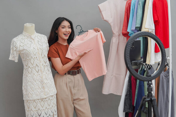 Attractive young Asian woman doing live stream online shopping pointing to the pink shirt she holds and presenting it to the camera. — Stock Image Attractive Young Asian Woman Doing Live Stream Online Shopping Pointing Stock Picture