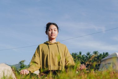 Calm young Asian woman is sitting in a field, wearing a green hoodie and doing yoga. She is in a peaceful and relaxed state, possibly meditating or enjoying the beauty of nature.