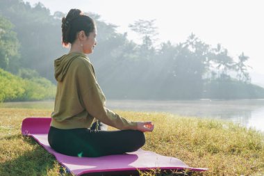 Side view of an Asian woman is sitting on a pink mat and doing yoga in a grassy field by the lake. She is wearing a green hoodie and black pants.