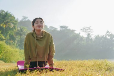 Focused Asian woman is doing yoga pose on a pink mat in a field. She is smiling and she is enjoying the outdoor activity.
