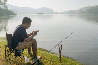 Young Asian man is sitting on a lawn chair by a lake, fishing and looking at his phone. The scene is peaceful and relaxing, with the man enjoying the outdoors and the calming sound of the water.