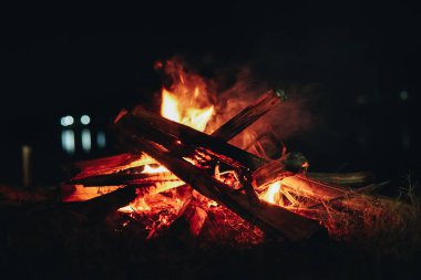 Ateş çukurunda bir odun yığını yanıyor. Sahne gecenin karanlığında çekiliyor, sadece ateşin ışığı bölgeyi aydınlatıyor..
