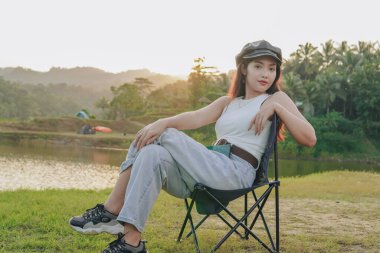 Zarif genç Asyalı kadın bir göl kenarında katlanır sandalyede oturuyor. Beyaz kolsuz bir bluz ve siyah bir şapka giyiyor..