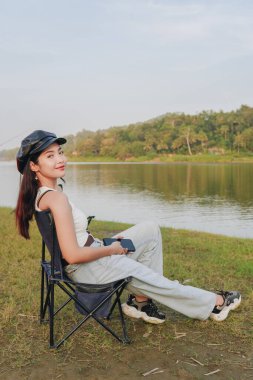 Şapka takan genç bir Asyalı kadının dikey portresi ve bir gölün kenarındaki sandalyeye oturmuş, Sermo Gölü, Yogyakarta, Endonezya manzarasının keyfini çıkarıyor..