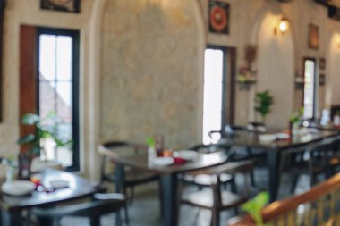 İçinde bir sürü masa ve sandalye olan bir restoranın portresi. Masalar vazo ve çiçeklerle dolu. Sandalyeler ahşap ve siyah. Ortam samimi ve davetkar..