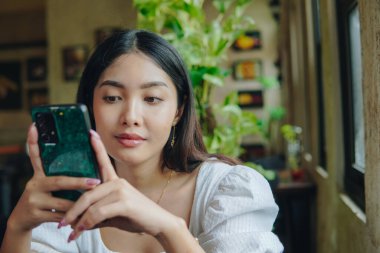 Beyaz bluz giyen, ciddi bir Asyalı kadın bir kafede otururken telefonuna sakin bir ifadeyle bakıyor..