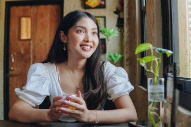 Güzel Asyalı kadın bir kafede oturmuş elinde bir bardak çayla kameraya gülümsüyor..
