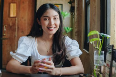 Güzel Asyalı kadın bir kafede oturmuş elinde bir bardak çayla kameraya gülümsüyor..