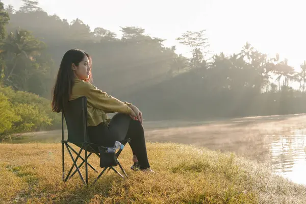 stock image Peaceful Asian woman sits in a lawn chair by a lake, looking out at the water. The scene is serene with the sun shining through the trees and casting a warm glow on the water.